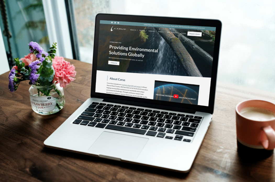 A laptop displaying a website about environmental solutions sits on a wooden table beside a flower vase with pink and purple flowers and a mug with a drink, near a window.